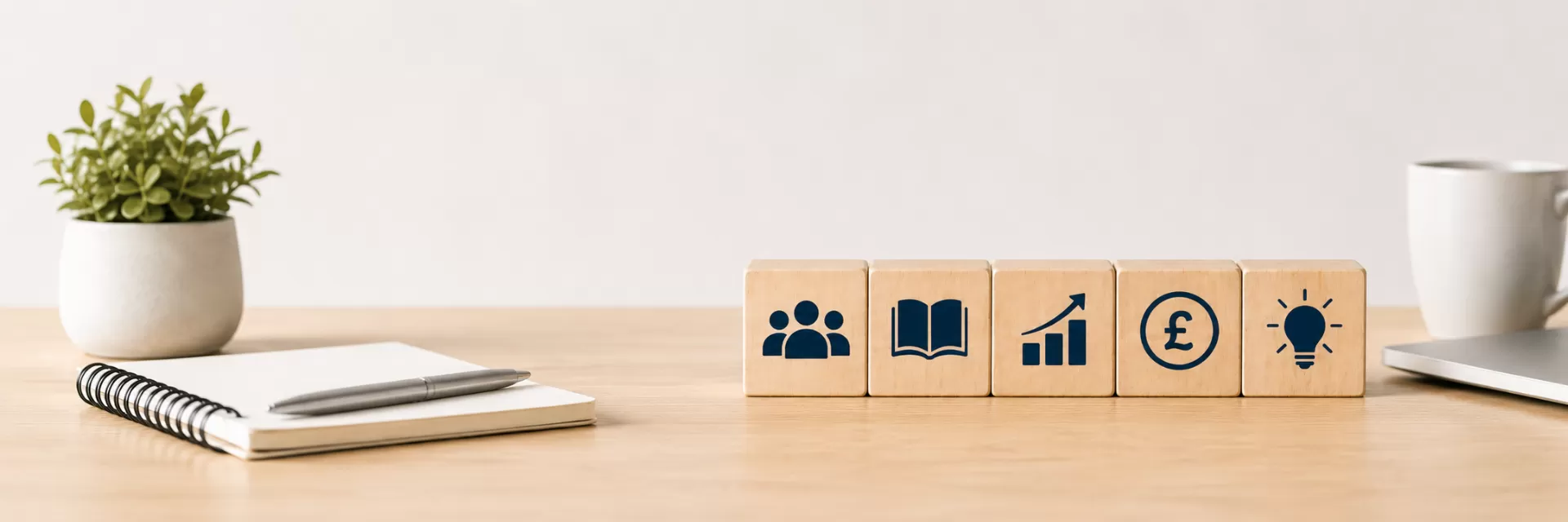 Minimal office desk scene with wooden blocks displaying icons representing people, learning, growth, finance and innovation, alongside a notebook, pen and plant in a modern workspace.